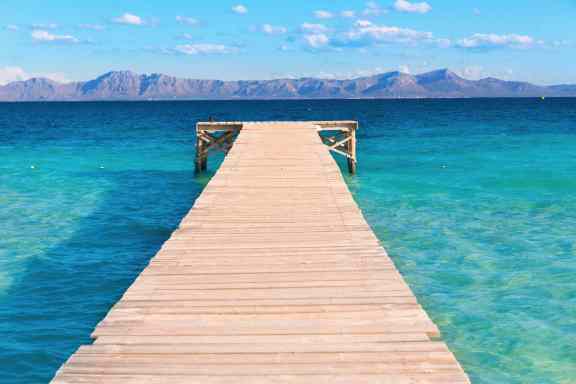 Long wooden pier in Alcudia, Mallorca stretching out into the clear blue water with a mountainous horizon.
